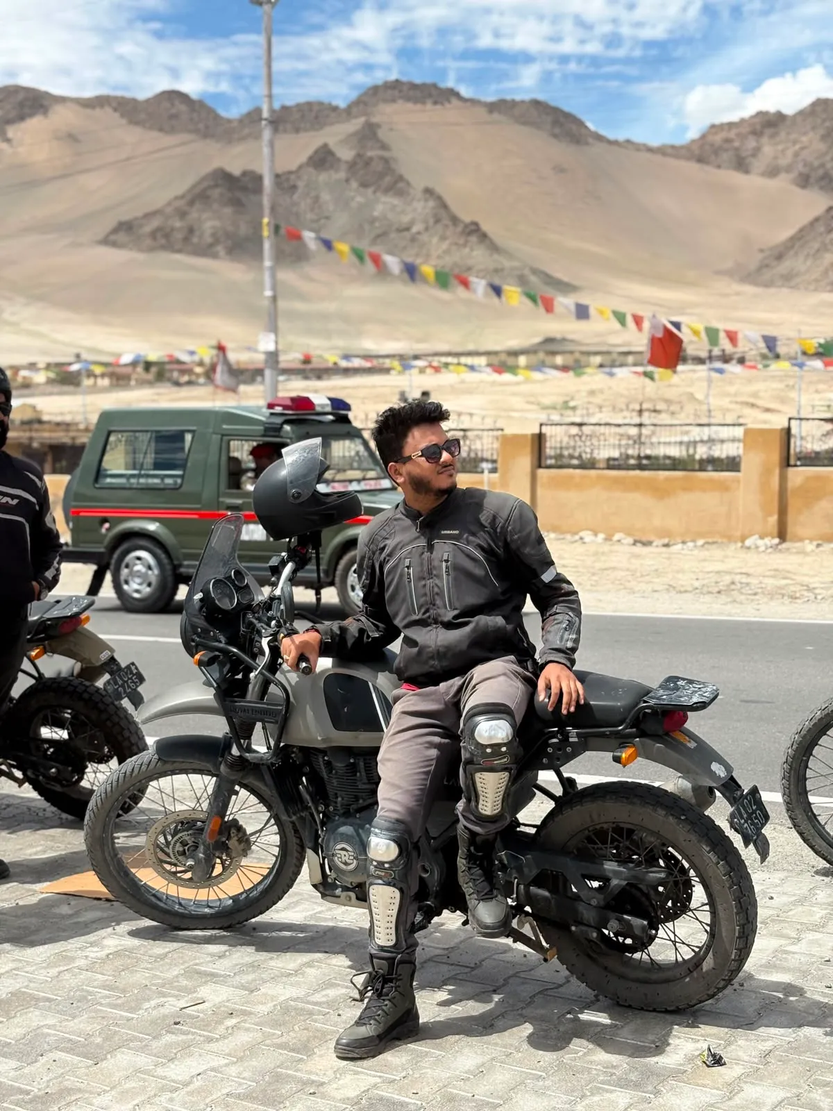 Debraj Roy in riding gear, resting on a Royal Enfield Himalayan on a Ladakh road with prayer flags and bare mountains behind him.