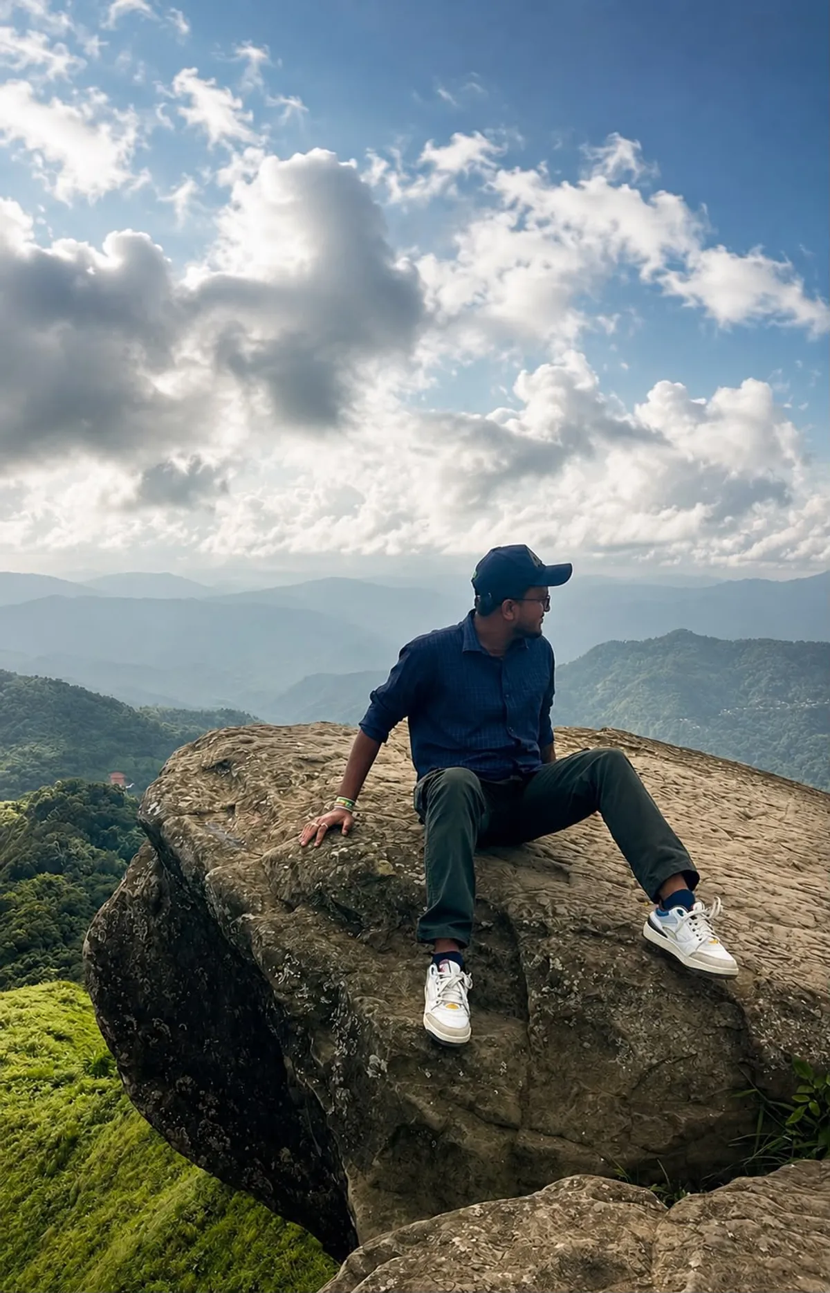 Debraj Roy sitting on a cliff edge, looking out over layered green hills and clouds.