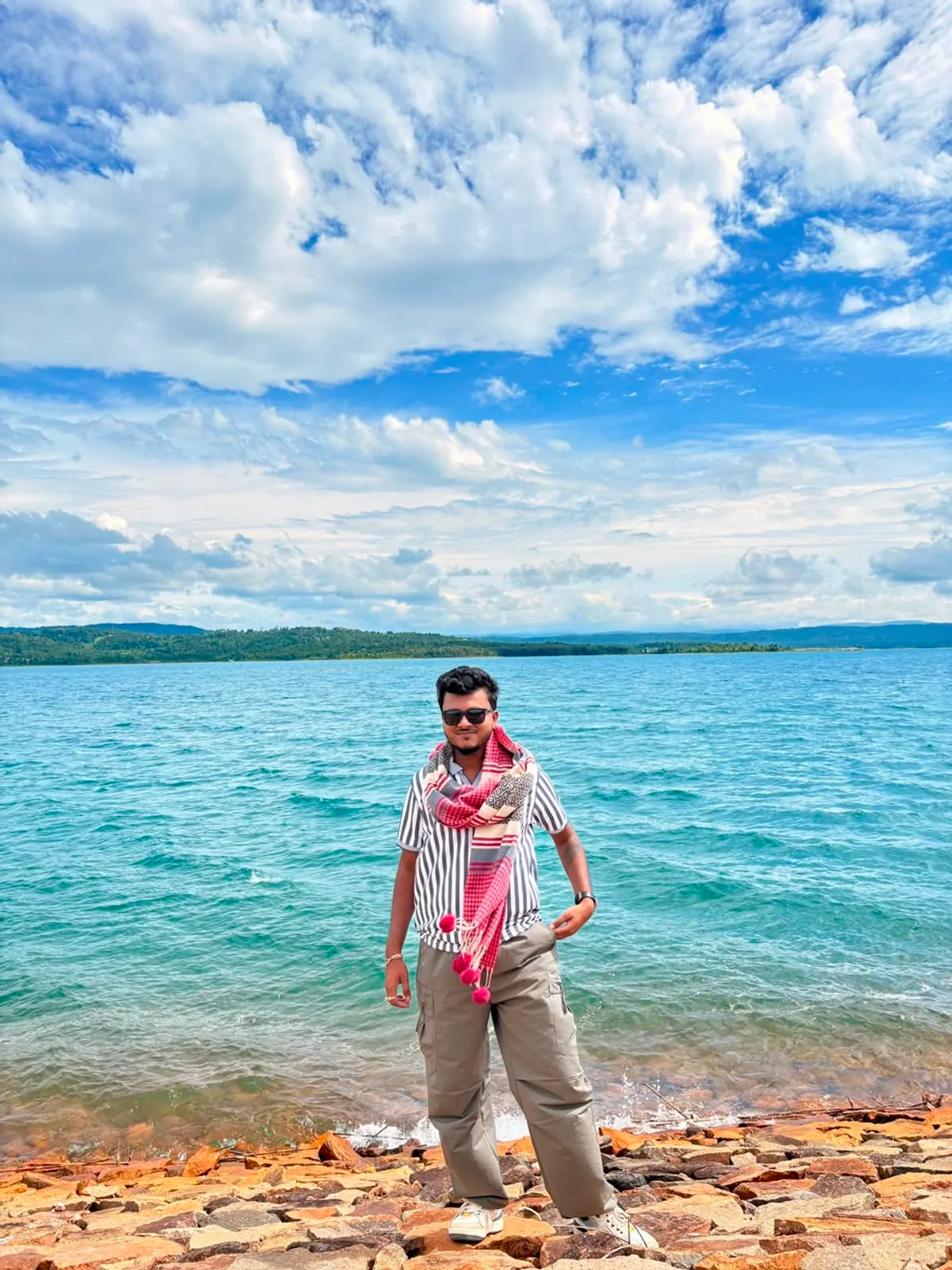 Debraj Roy standing at a lakeshore in Assam, wearing a traditional gamusa around his neck.